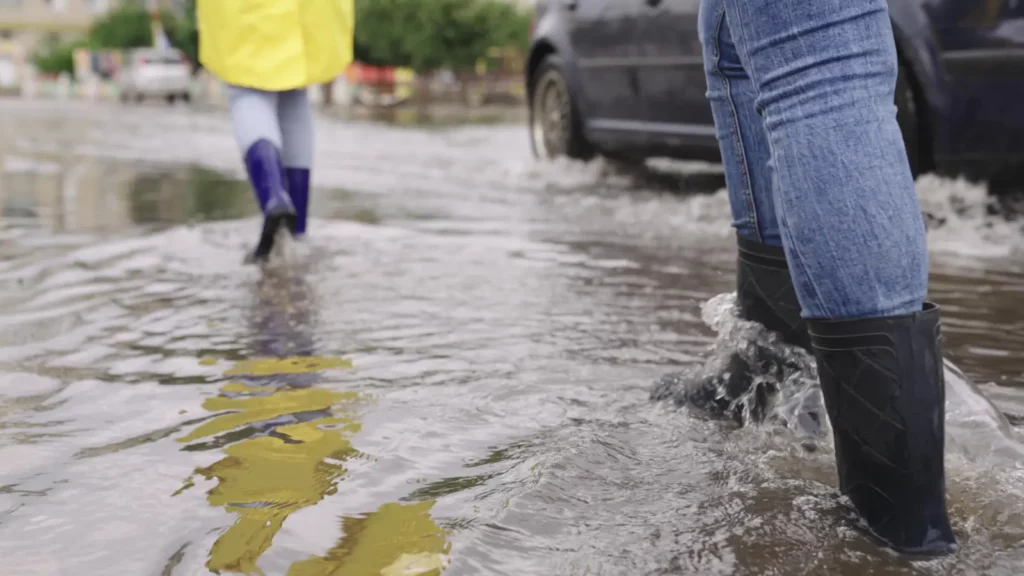 Two people wearing boots walk down a flooded street. Only their legs are shown.