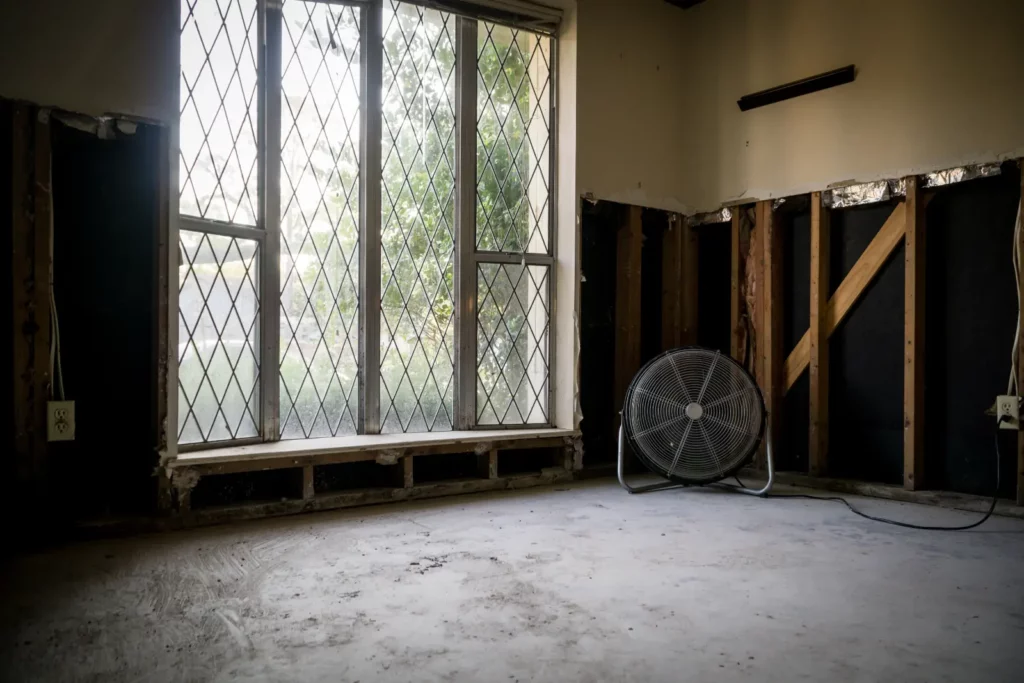 A room with flood damage. The walls have been stripped to the studs and a large fan is in the corner.